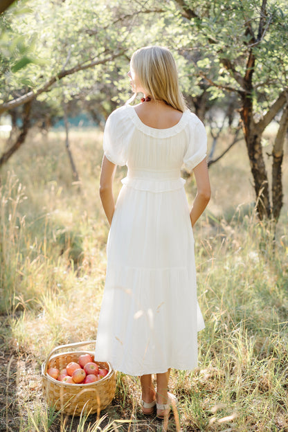 Back view of a woman wearing a white midi dress with a scoop necklien, puff sleeves and wide bottom ruffle.