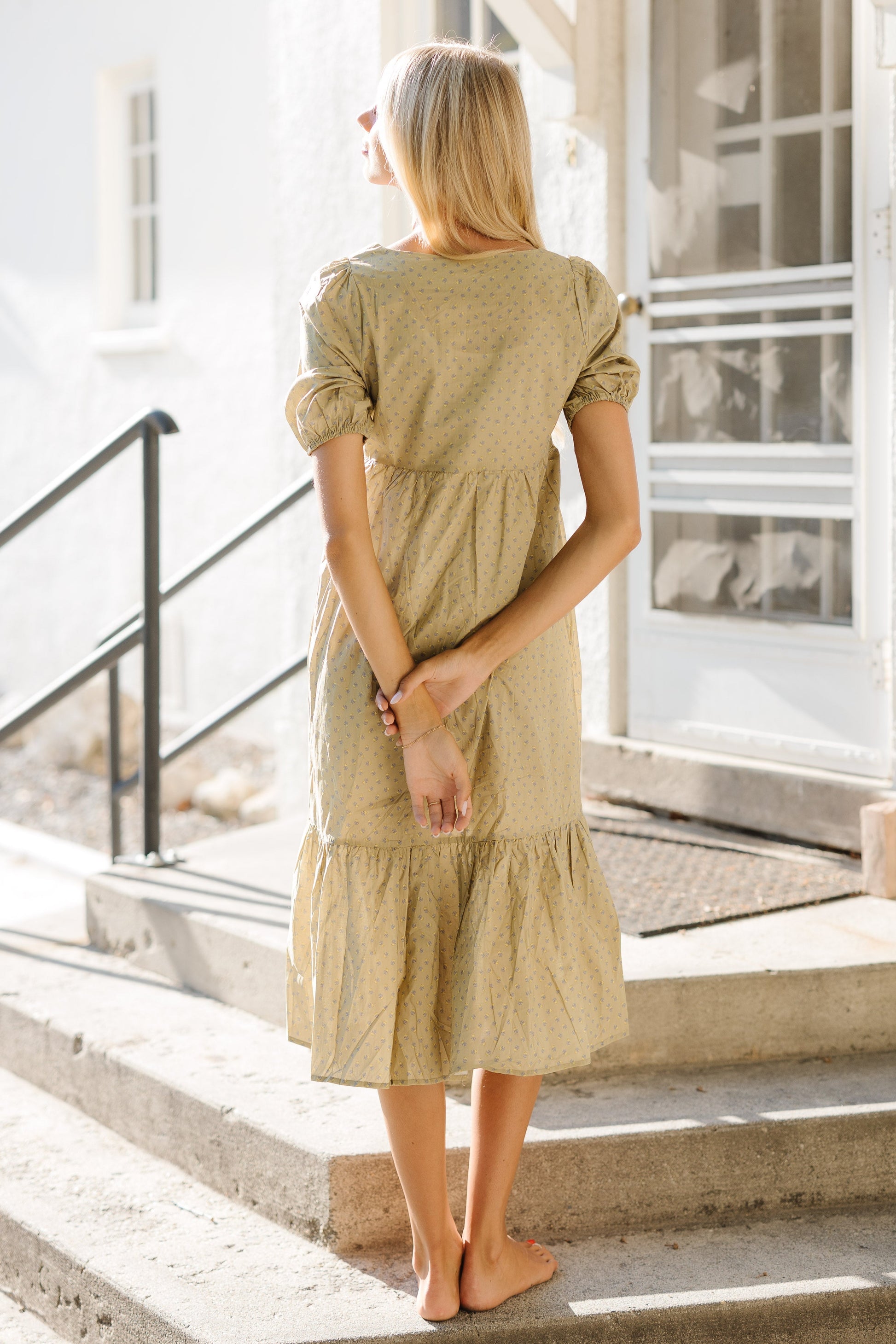 Back view of a young woman wearing an olive green print midi dress with short puff sleeves and a wide bottom ruffle.