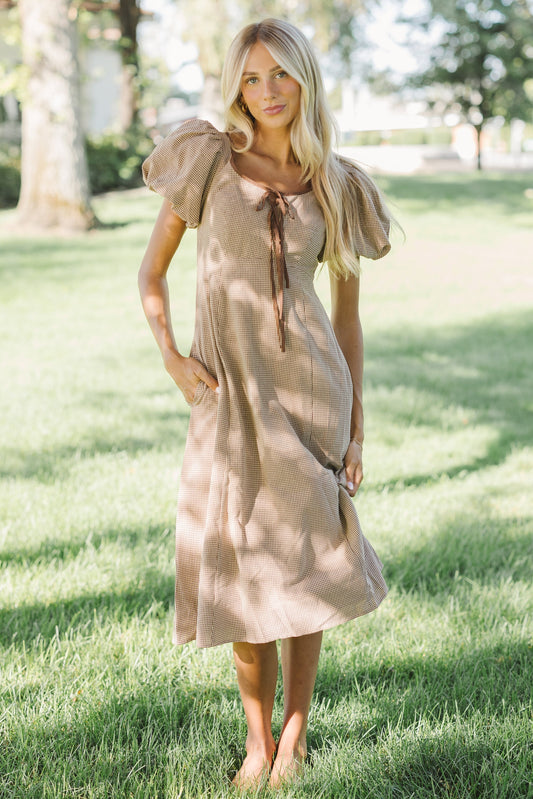 Young womna wearing a modest brown and white gingham dress with a wide scoop neckline, balloon sleeves, A-line skirt & pockets.