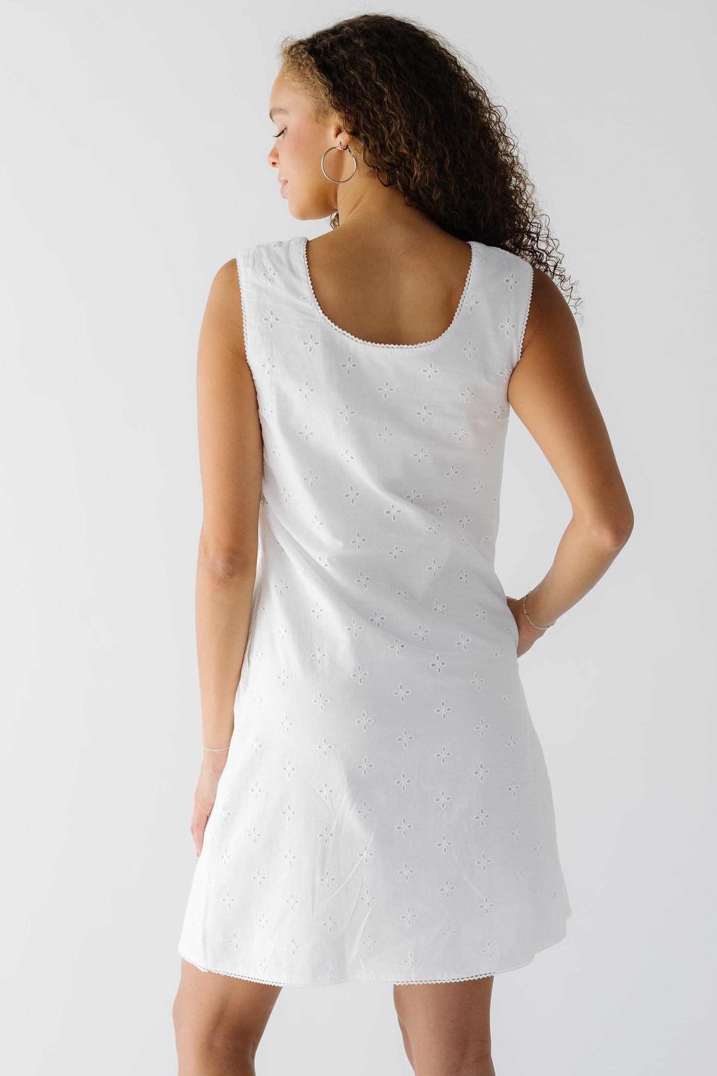 Back view of a woman wearing  a sleeveless white eyelet shift dress with a back scoop neckline.