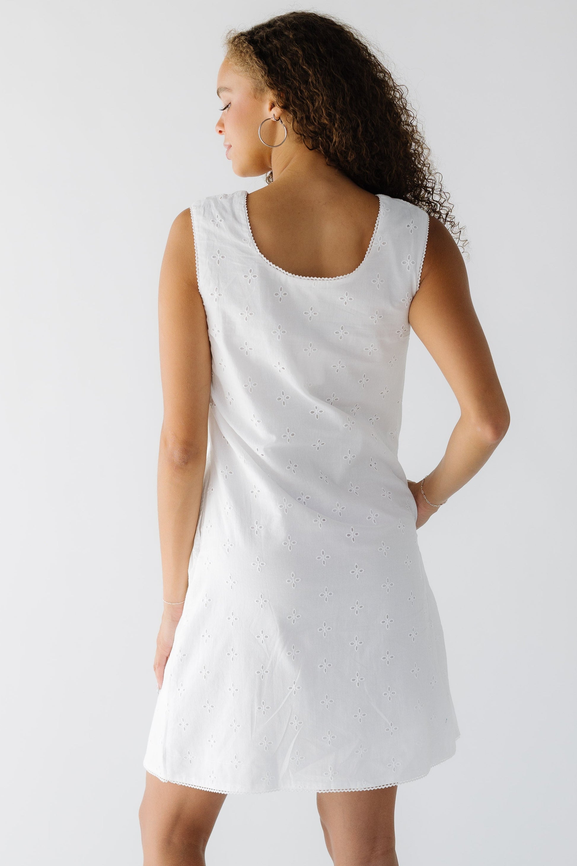 Back view of a woman wearing  a sleeveless white eyelet shift dress with a back scoop neckline.