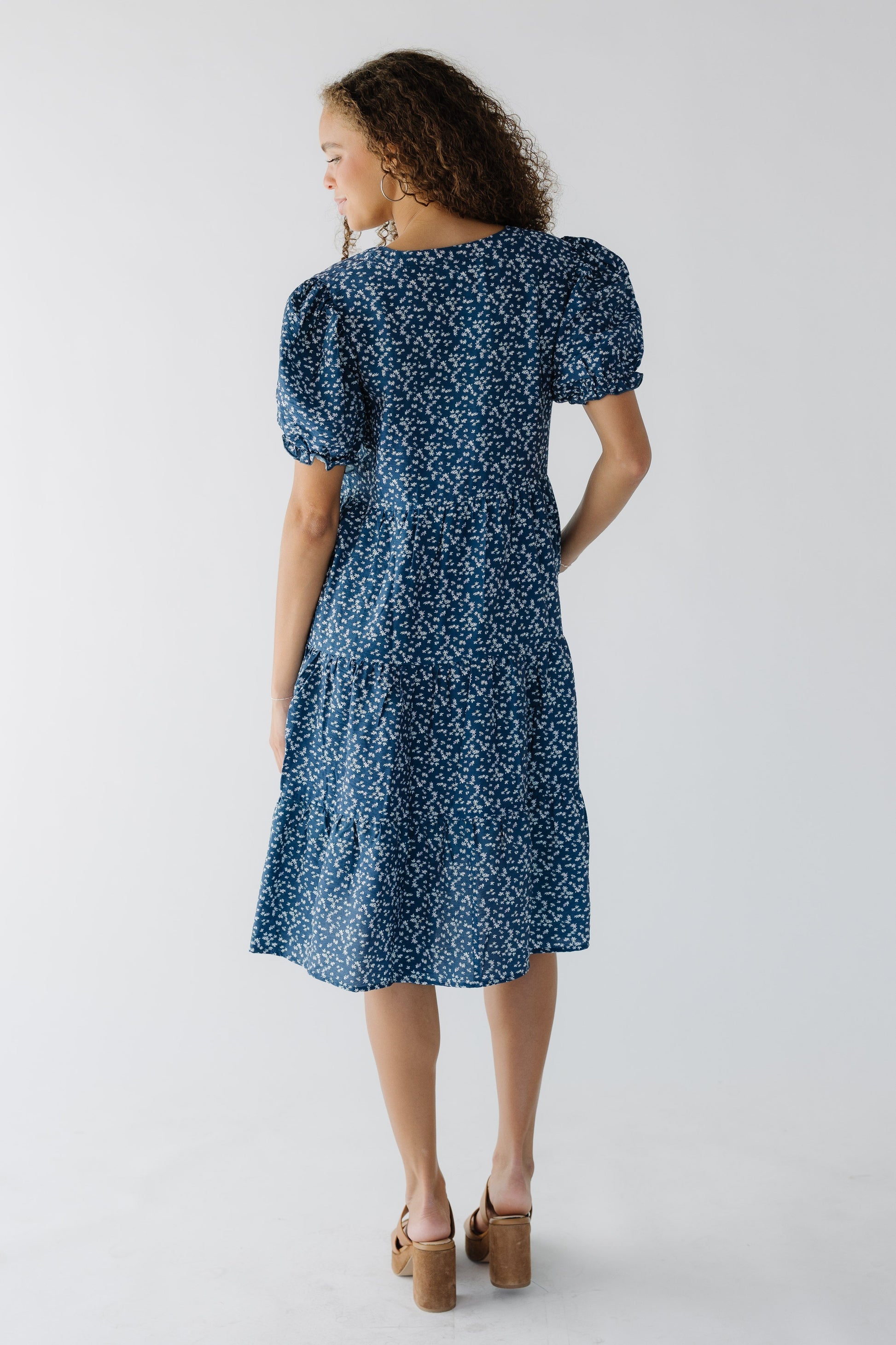 Back view of a woman wearing a knee length blue floral tiered dress with short puff sleeves.