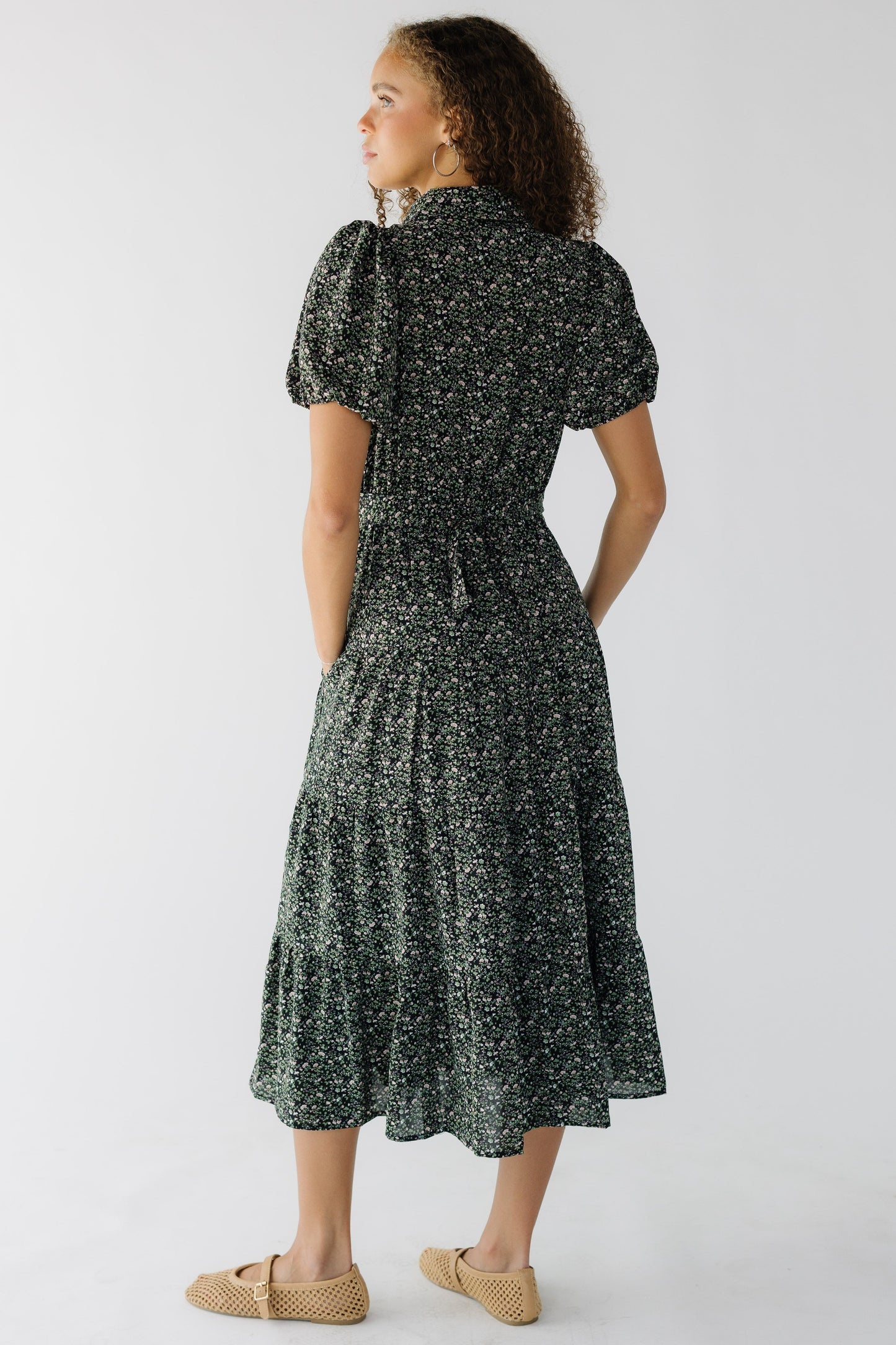Back view of a teenage girl weaing a black floral, tiered midi dress wtih short sleve and a clser fitting waist.