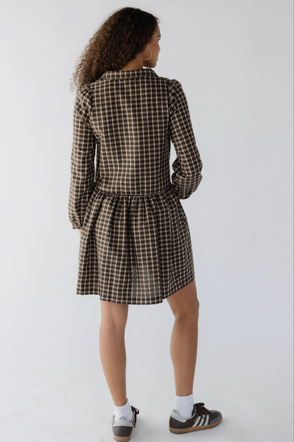Back view of a teen age girl wearing a short brown plaid dress with long sleeves.