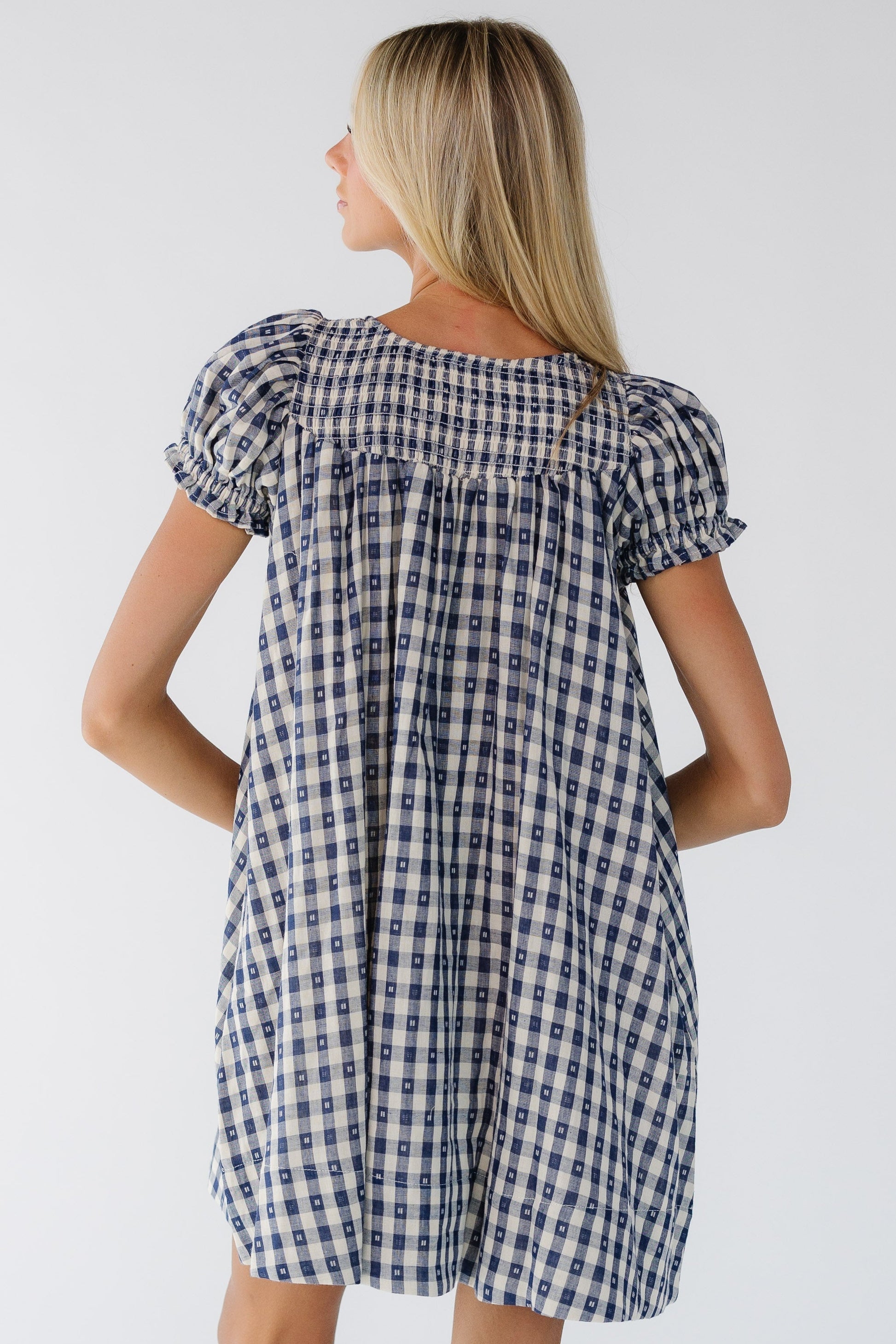 Back view of a woman wearing a navy & whie gingham knee dress with short puff sleeves and a smocked back neckline.