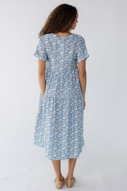 Back view of a teen girl wearing an ivory and blue floral midi dress with side pockets and short sleeves.
