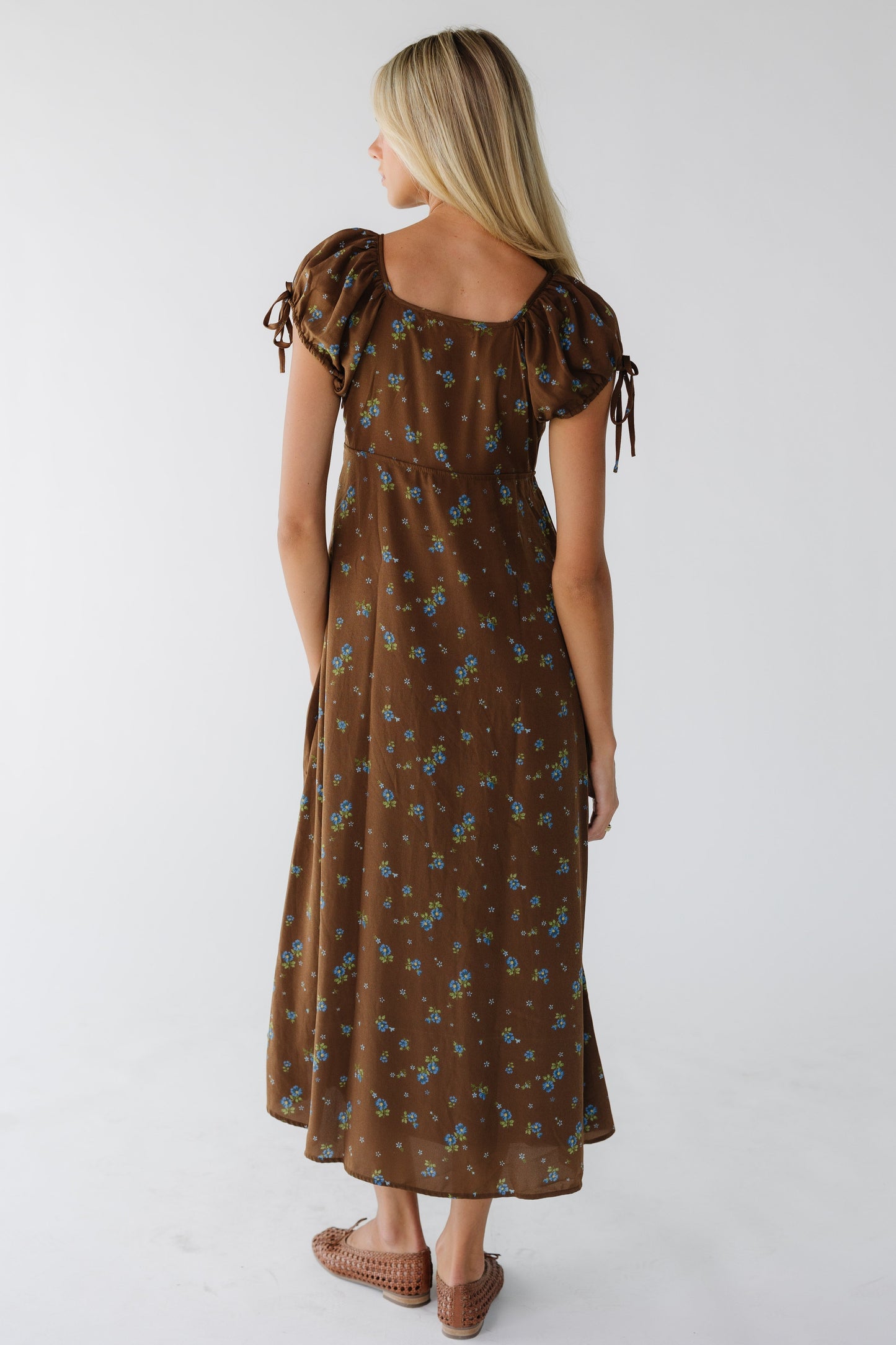 Back view of a young woman wearing a brown & blue floral midi maxi dress with a square neckline and puff sleeves.