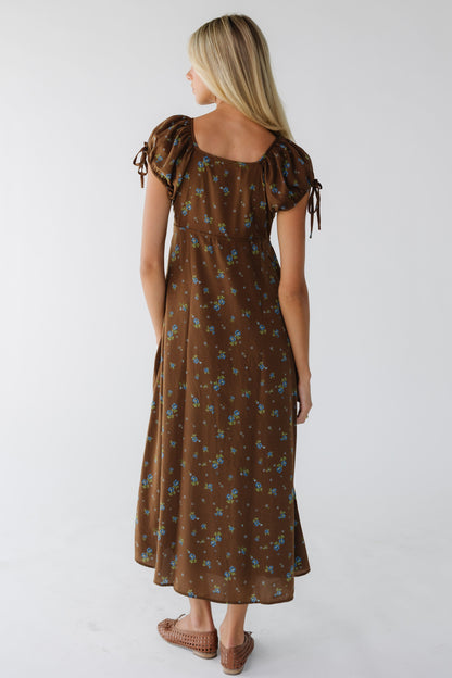 Back view of a young woman wearing a brown & blue floral midi maxi dress with a square neckline and puff sleeves.