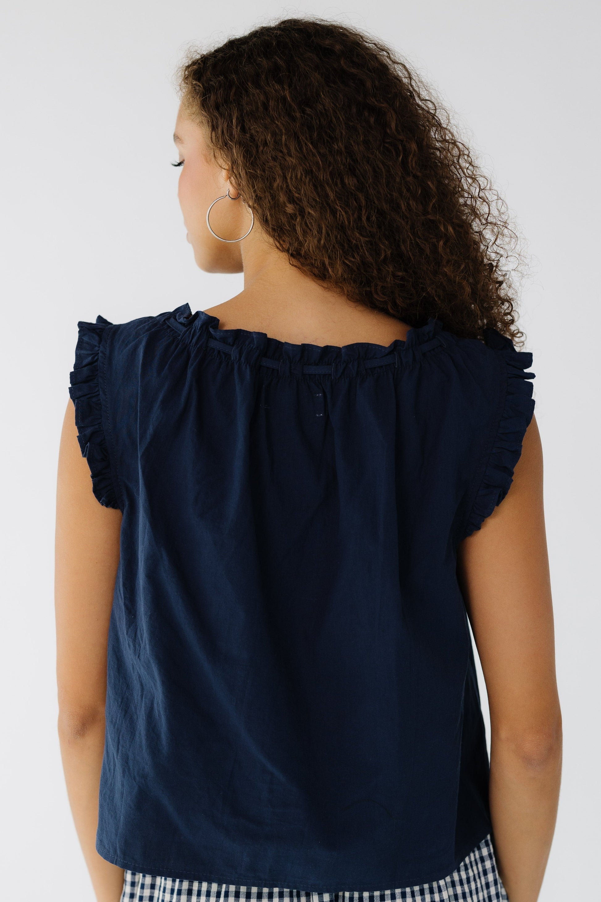 Back view of a teen girls wearing a navy loose fitting, sleeveless blouse with ruffles around the neckline and arm openings.
