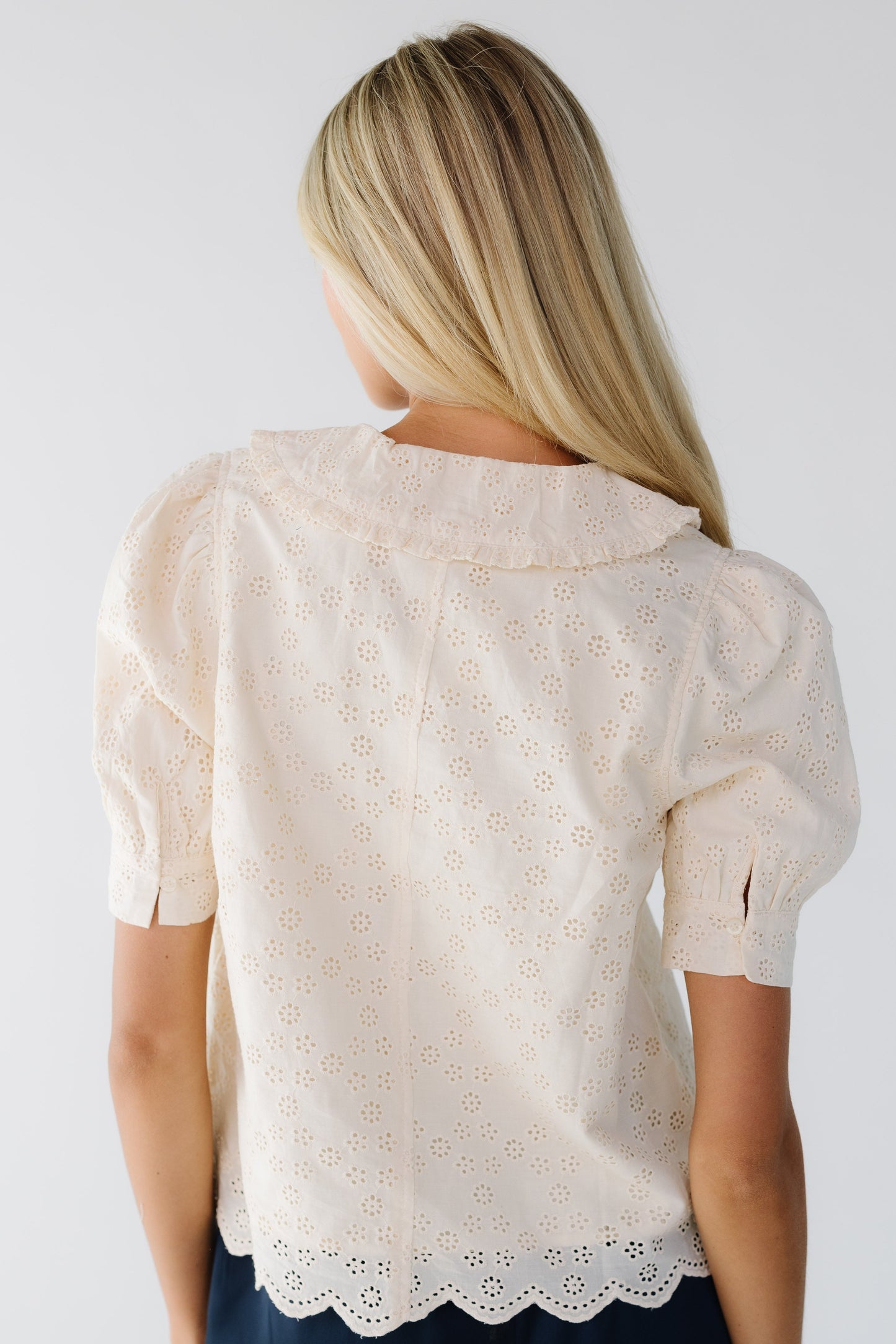 Back view of a young woman wearing a lined, cream eyelet blouse with short cuffed sleeves and a ruffle edged collar.