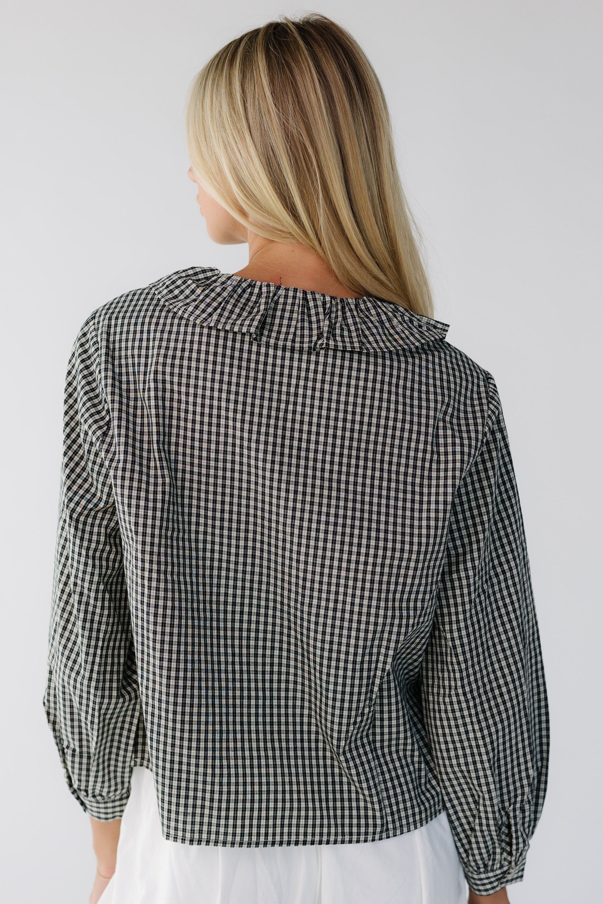 Back view of a woman wearing a black plaid loose fitting blouse with long sleeves and a ruffled collar.