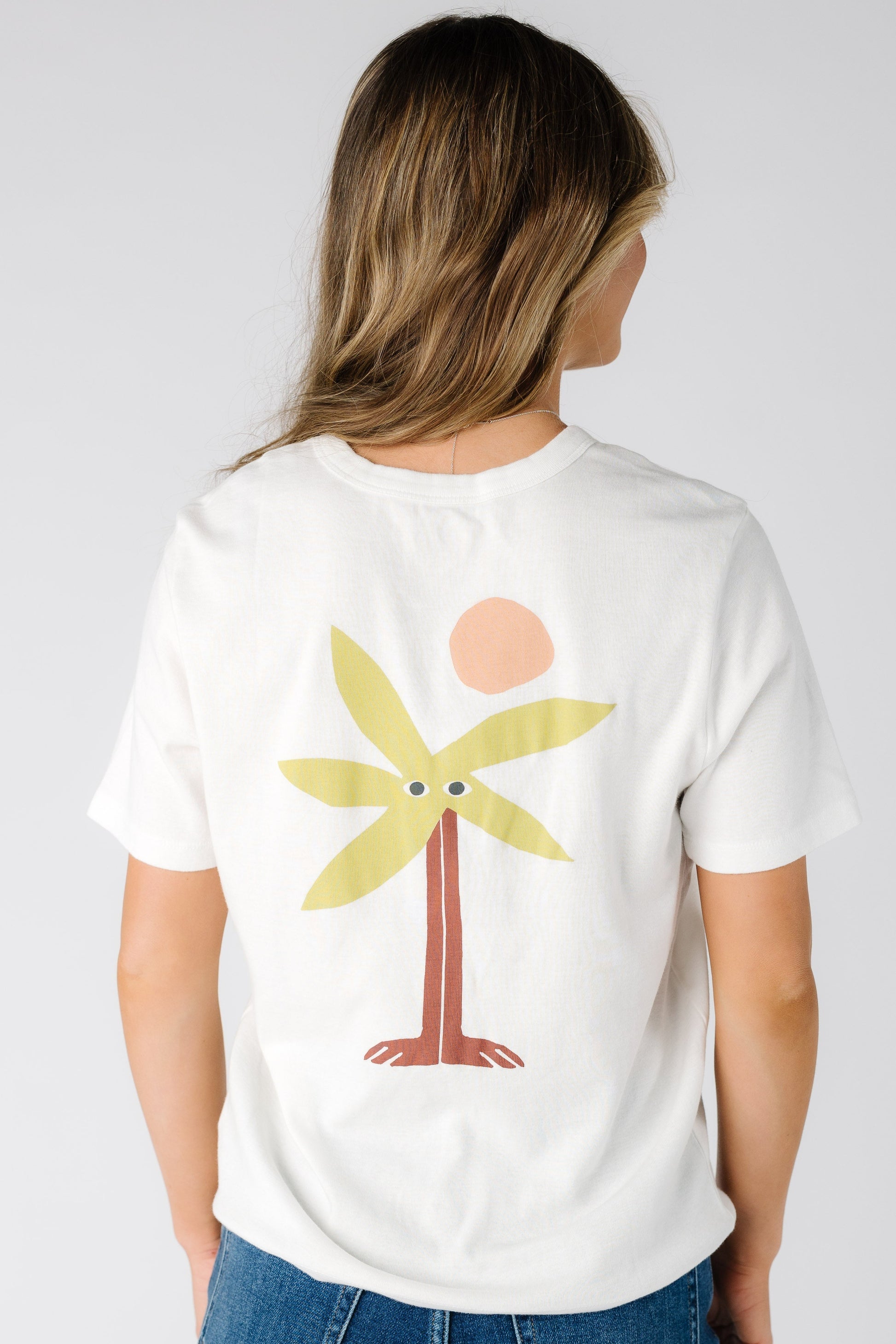 Back view of a woman wearing a white graphic tee with a large back graphic of  a palm tee and sun.