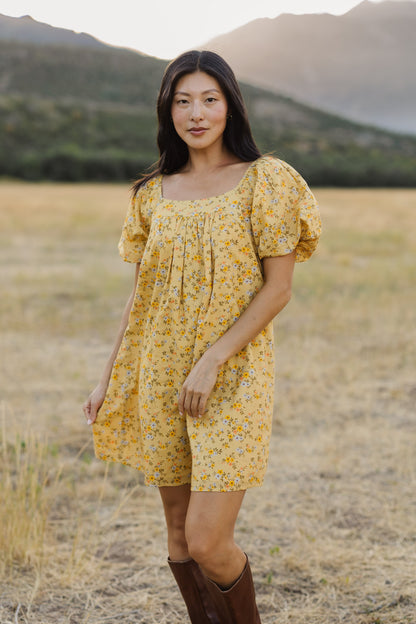 A young woman wearing a short yellow, floral print dress with a square neckline and balloon sleeves.