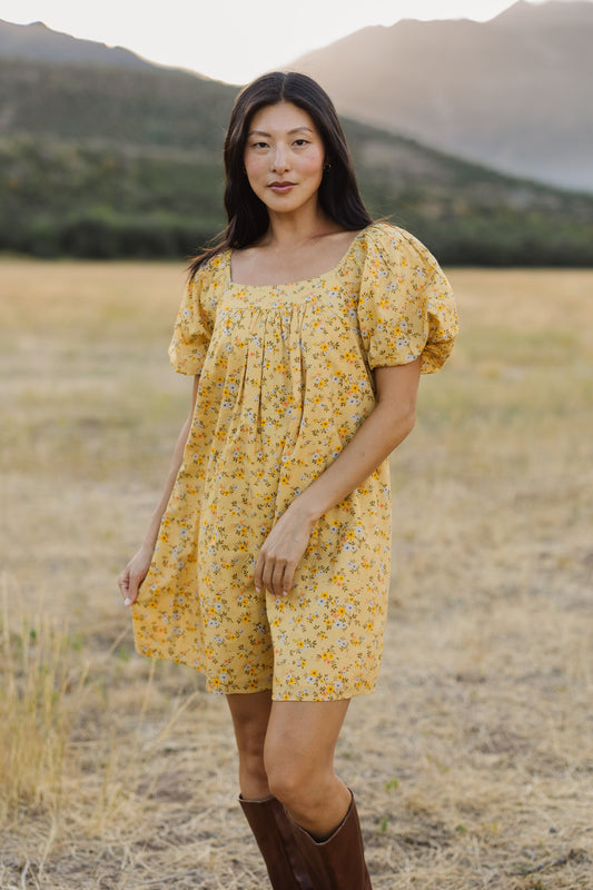 A young woman wearing a short yellow, floral print dress with a square neckline and balloon sleeves.