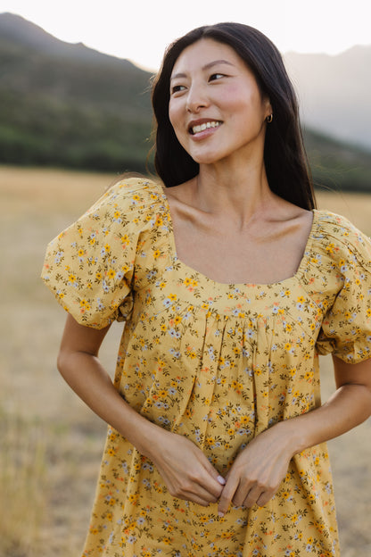 A close up view of a woman wearing a yellow floral print dress with a square neckline and balloon sleeves.