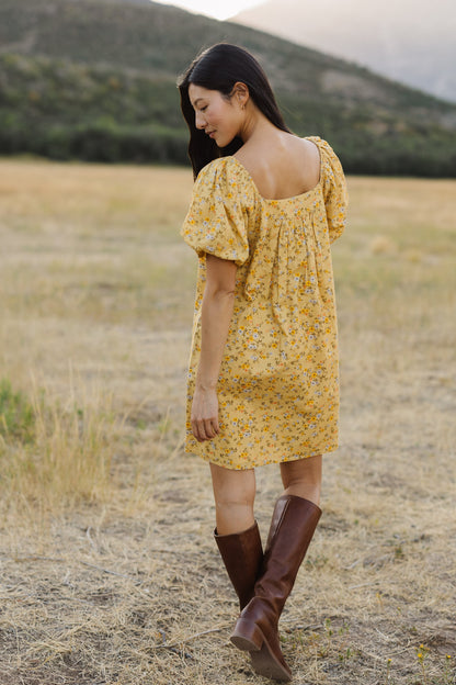 Back view of a young woman wearing a short yellow floral print dress with a square neckline and balloon sleeves.