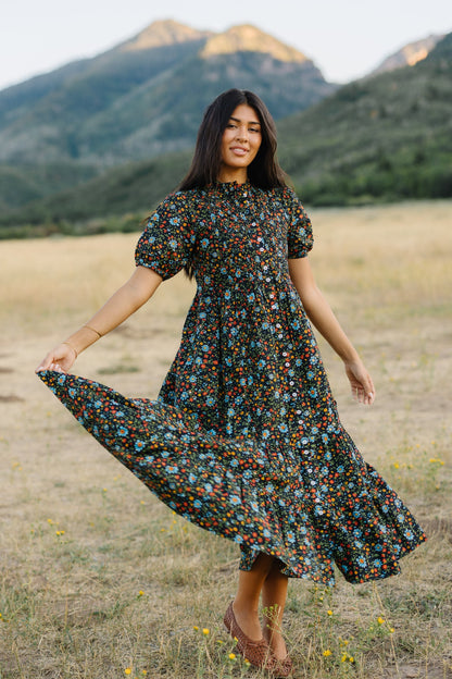 Young woman wearing a black floral print midi-maxi, button down dress with short puff sleeves, ruching and pleats and a wide botto ruffle.
