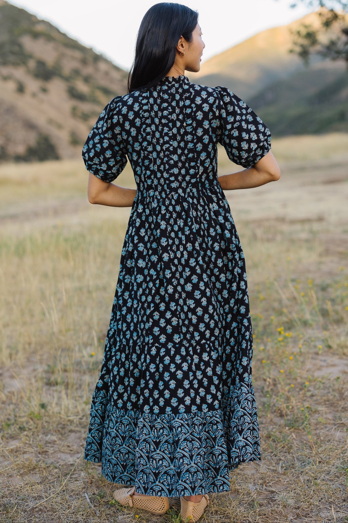 Back view of a woman wearing a navy & blue floral midi-maxi dress with a wide bottom ruffle in a different floral print.