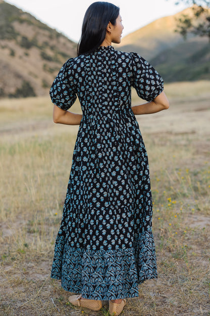 Back view of a woman wearing a navy & blue floral midi-maxi dress with a wide bottom ruffle in a different floral print.