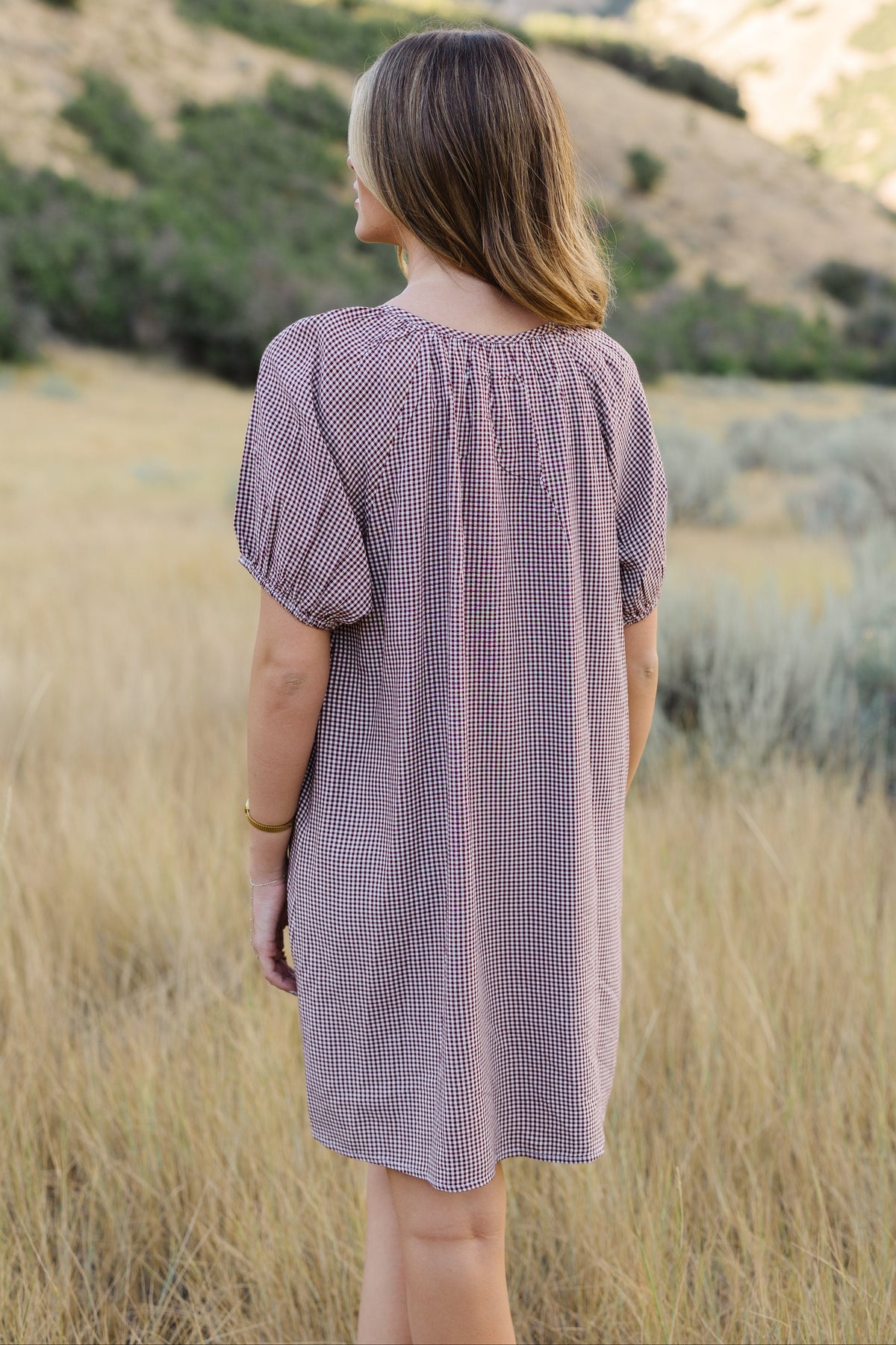 Back view of a woman wearing a short burgundy & white gingham dress with large puff sleeve.
