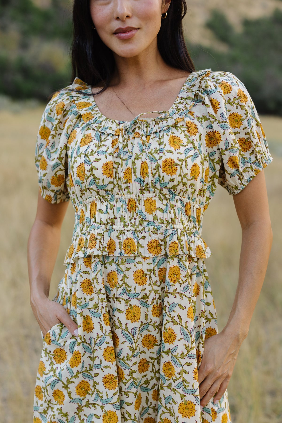 Close view of a woman wearing a floral print dress with a scoop neckline with a ruffle, short puff sleeves, and a wide waistband and full skirt.