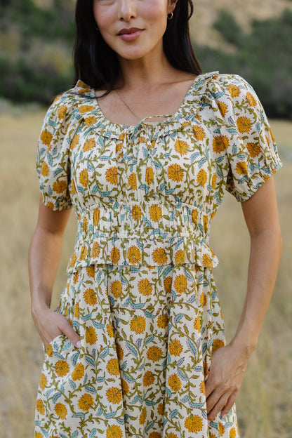 Close view of a woman wearing a floral print dress with a scoop neckline with a ruffle, short puff sleeves, and a wide waistband and full skirt.