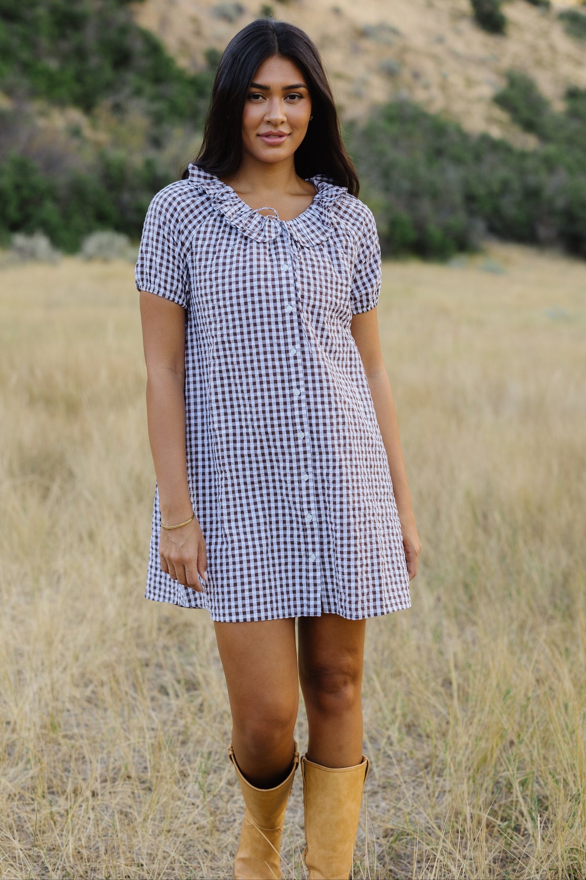 Young woman wearing a brown & white gingham, button down short dress with a round ruffle neckline and puff sleeves.
