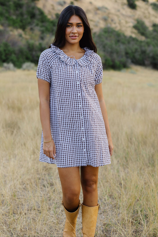 Young woman wearing a brown & white gingham, button down short dress with a round ruffle neckline and puff sleeves.