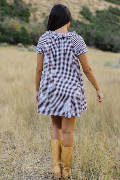 Back view of a woman wearing a brown & white gingham short dress with short puff sleeves and ruffed collar.