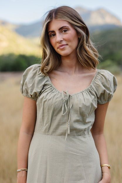 Close up view of a woman wearing a a light green gingham dress with a drawstring scoop neckline with puff sleeves.
