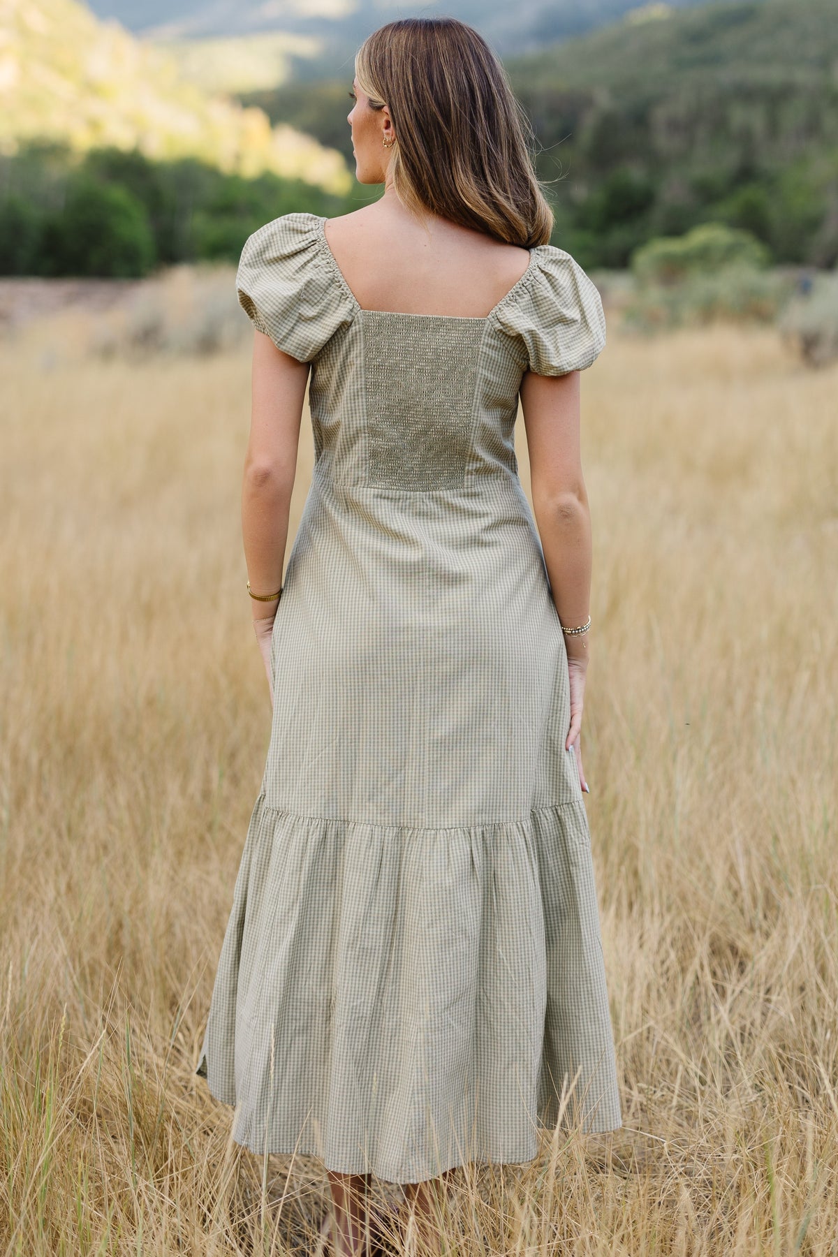 Back view of a woman wearing a light green gingham midi dress with an elastic smocked back bodice, square neckline and short puff sleeves.