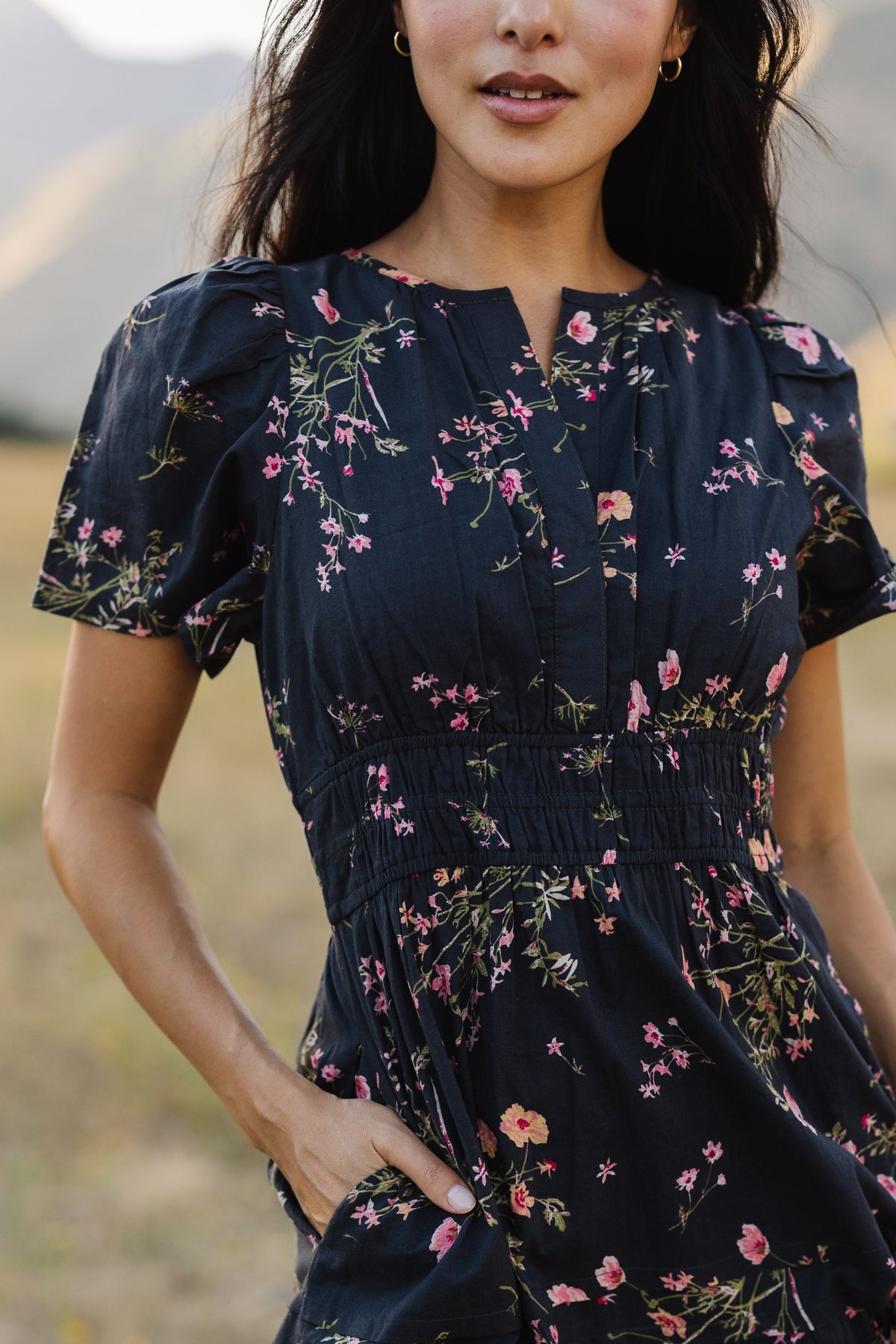 Close up view of a woman wearing a navy floral print dress with a V-neckline, short flutter sleeves, wide elastic waistband and side pockets.