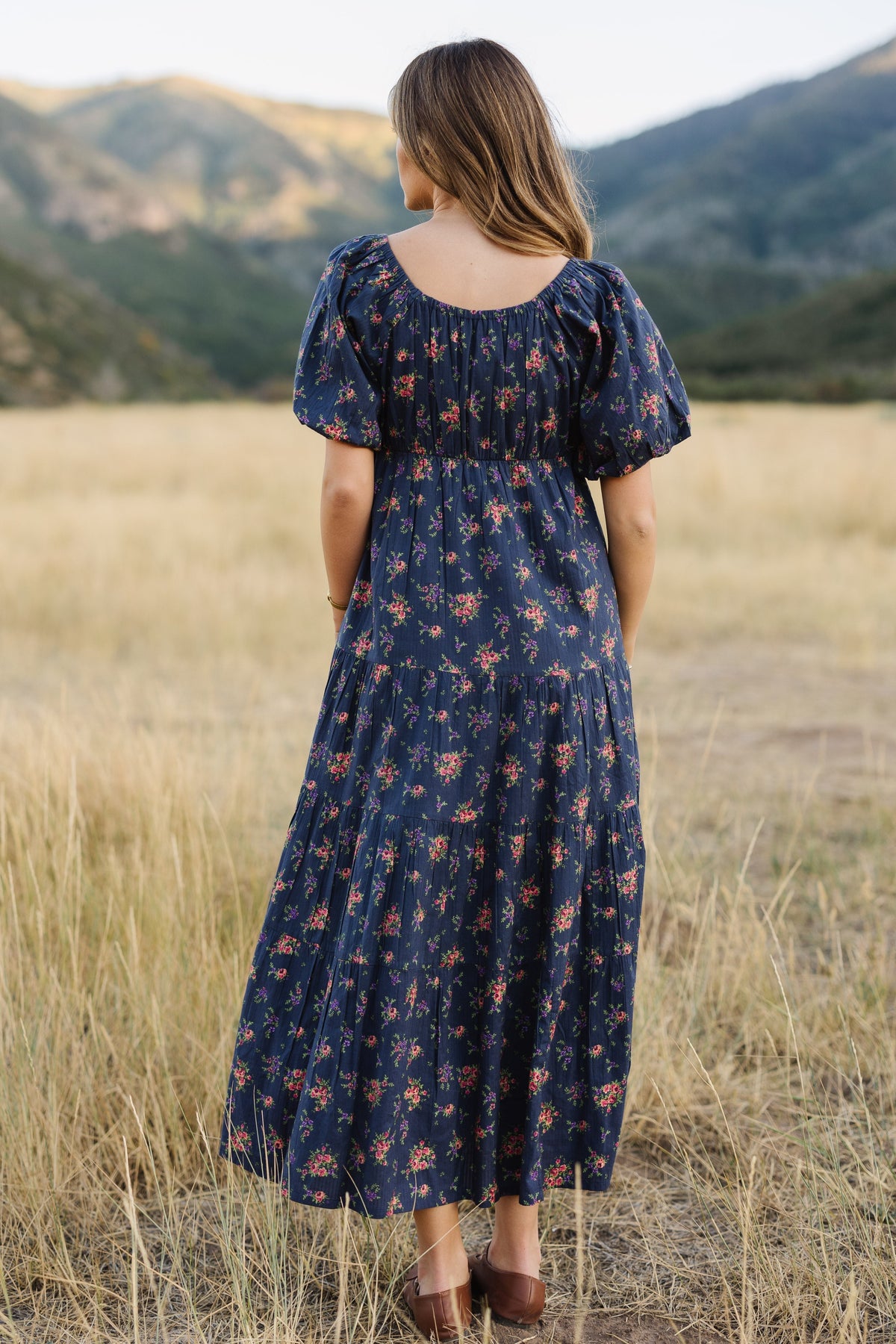A back view of a woman wearing a navy floral midi dress with arge puff sleeves and a bakc scoop neckline.