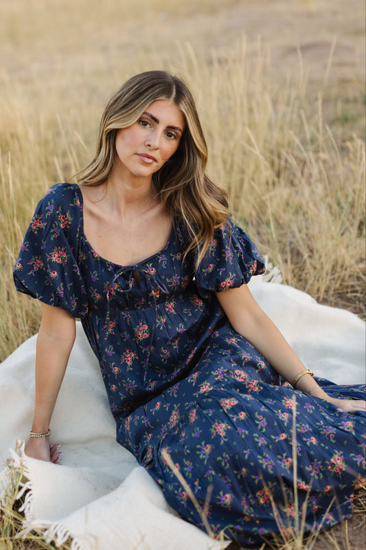 A young woman wearing a navy floral midi dress with a scoop necklie, large puff sleeves and an empire waist.