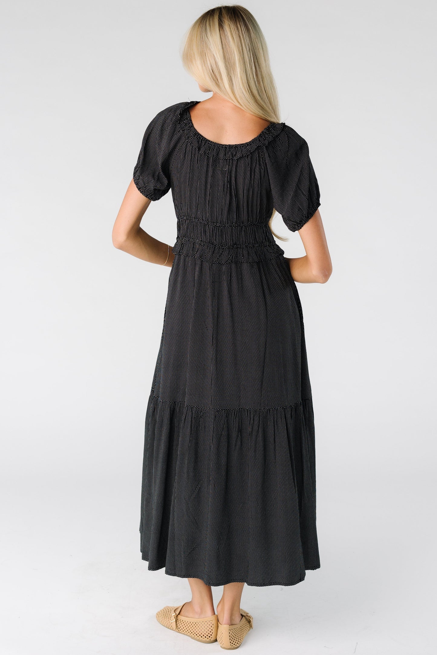 Back view of a woman wearing a black polka dot midi dress with a wide bottom ruffle, short balloon sleeves and a wide waist and scoop neckline.