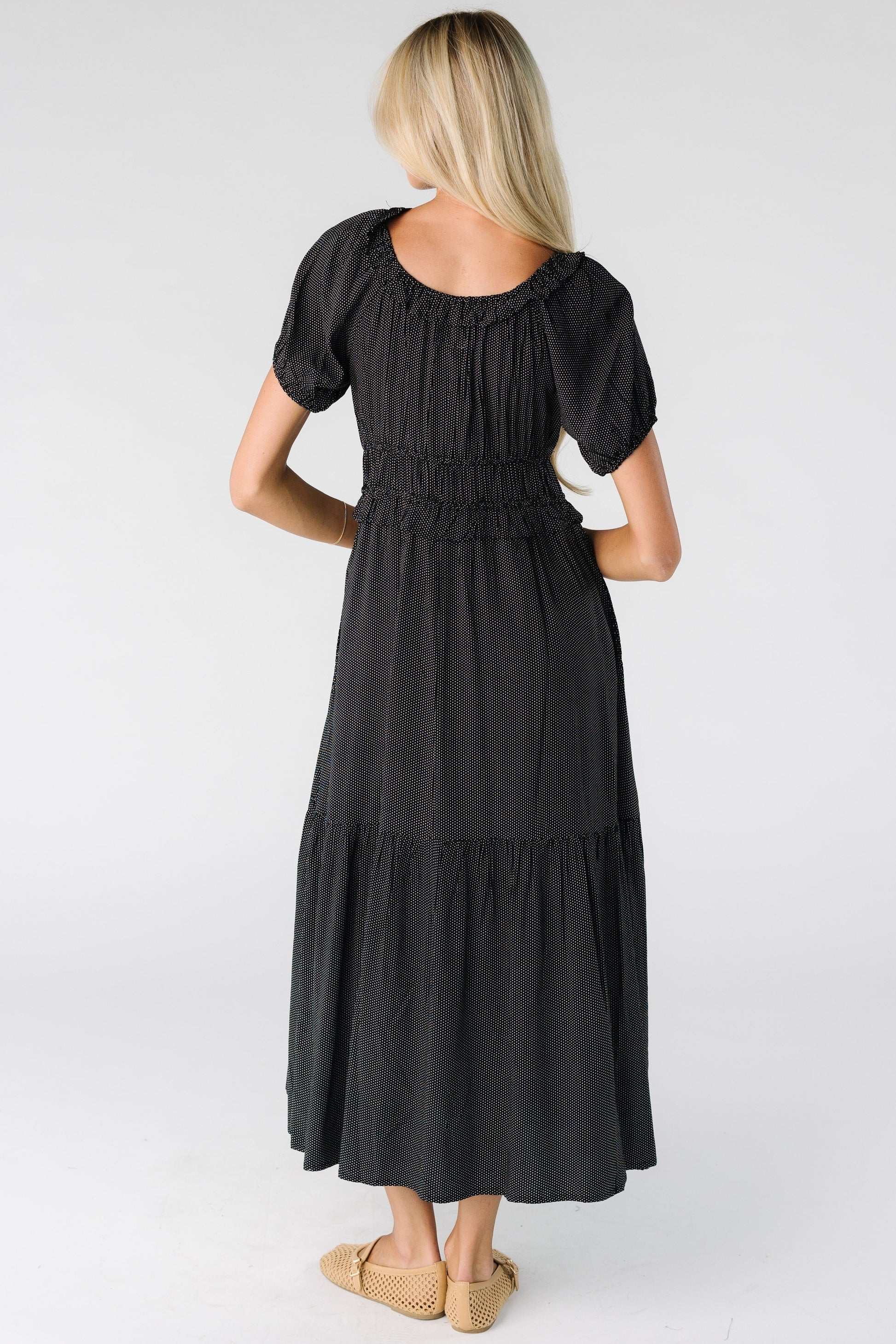 Back view of a woman wearing a black polka dot midi dress with a wide bottom ruffle, short balloon sleeves and a wide waist and scoop neckline.