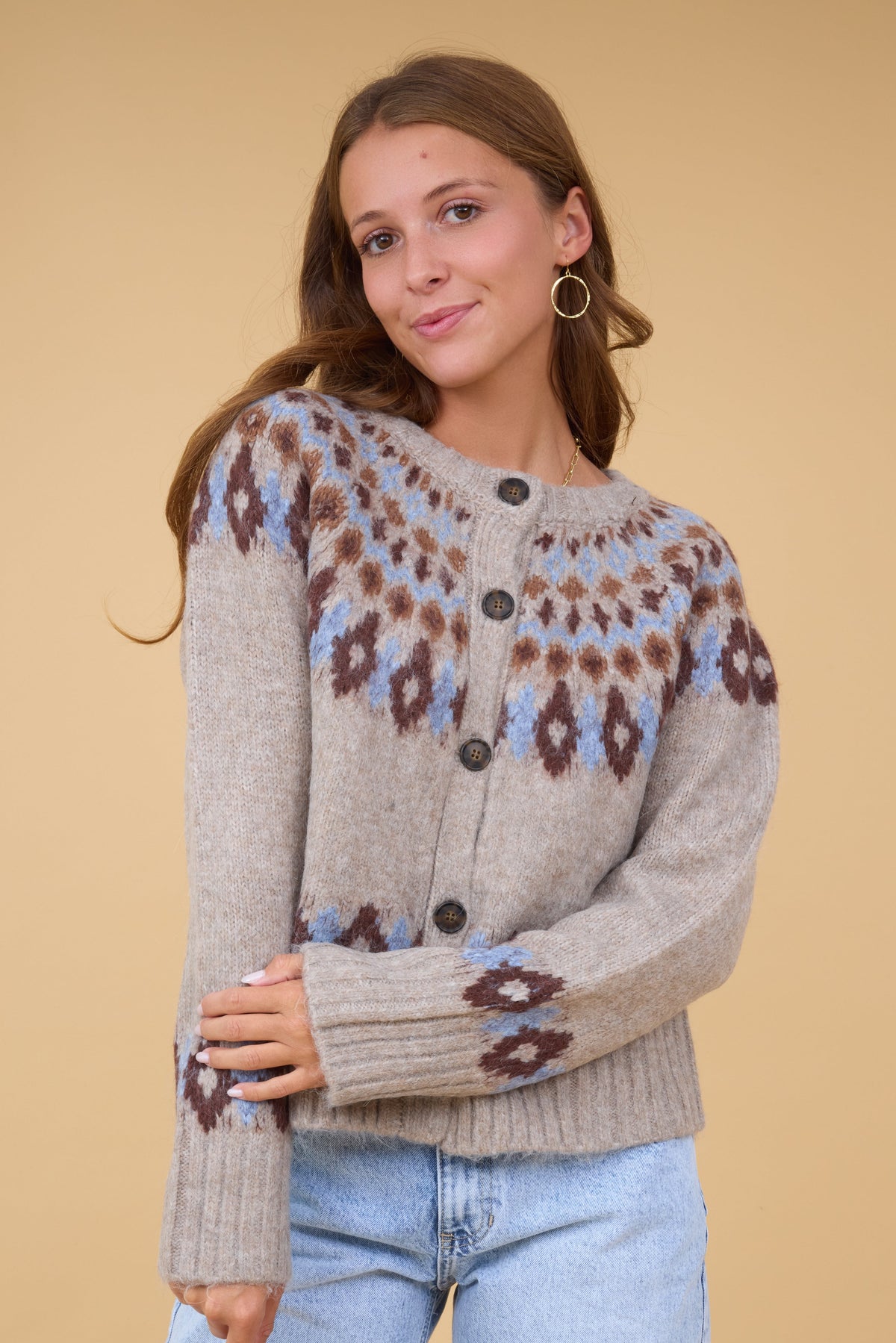 A young woman wearing a modest tan cardigan with large buttons with a blue & brown nordic pattern.