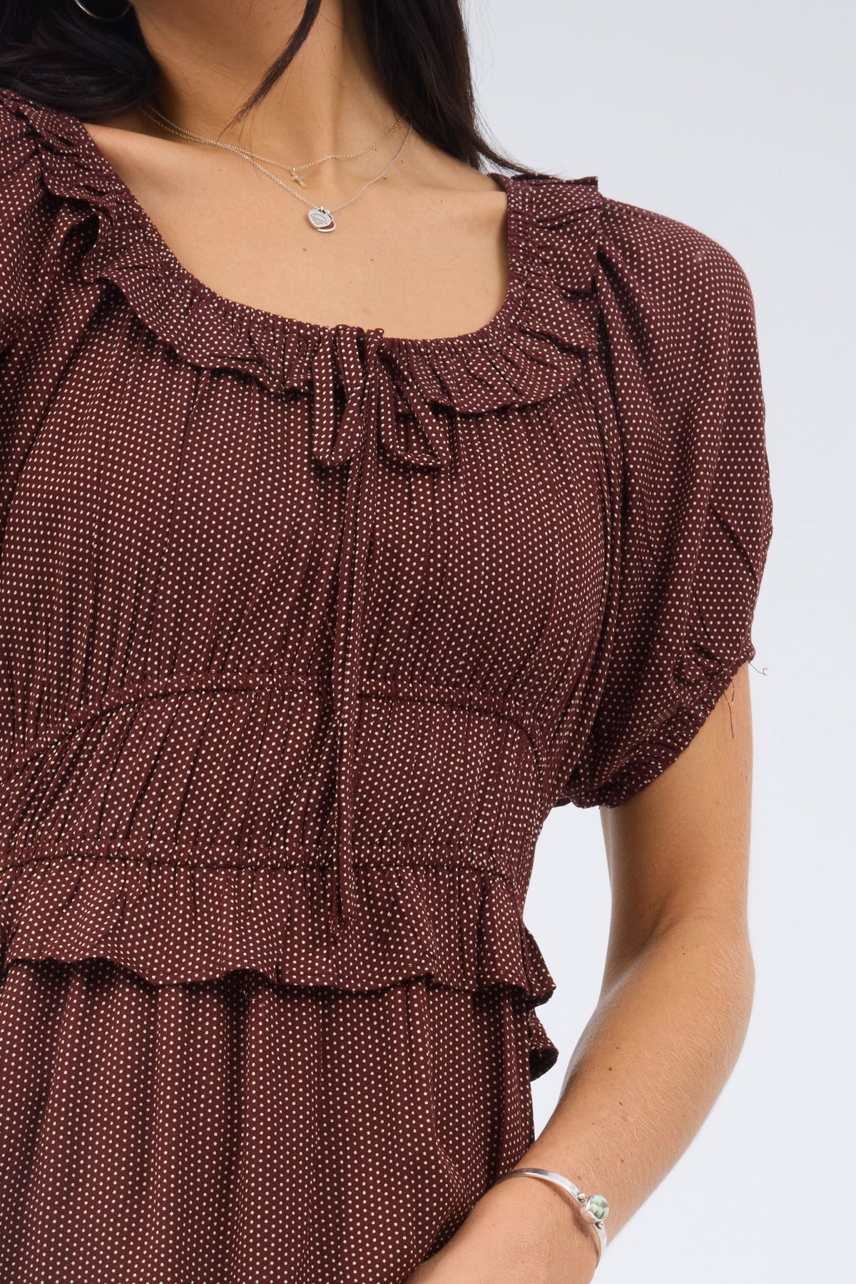 A close up view of a woman wearing a brown and white polka dot dress with a ruffled scoop neckline, large puff sleeves and a wide wiastband and a  narrow ruffle.