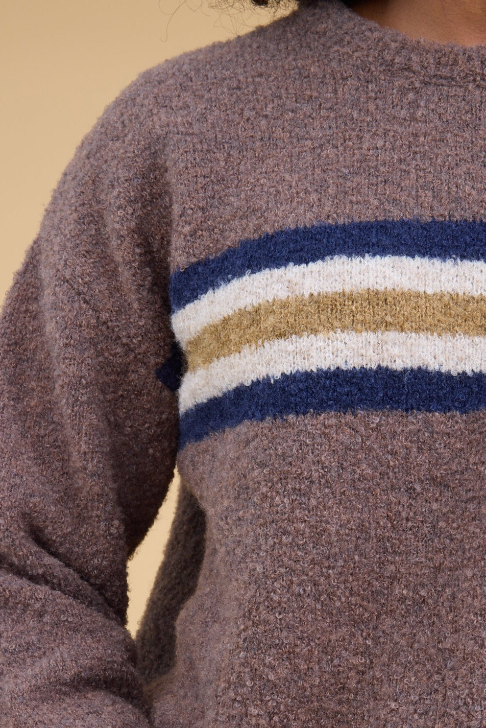 A close up view of a woman wearing a light brown slubby sweater with a wide stripe around the chestin blue, white & gold.
