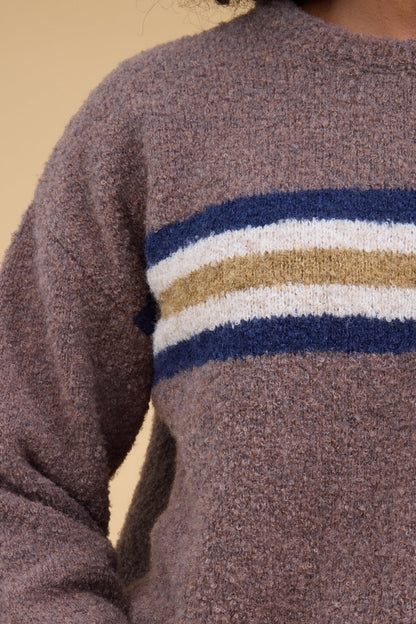 A close up view of a woman wearing a light brown slubby sweater with a wide stripe around the chestin blue, white & gold.