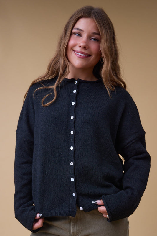 Young woman wearing a black cardigan with white buttons, long sleeves and a rolled neckline.