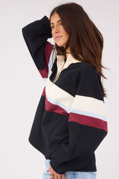 A side view of a woman wearing a loose fitting black pullover top with long sleeves and wide stripe in cream, light blue & maroon.