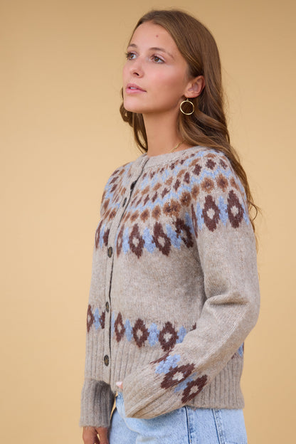 A woman wearing a beige buttond down cardigan wtih a blue & brown nordic pattern.