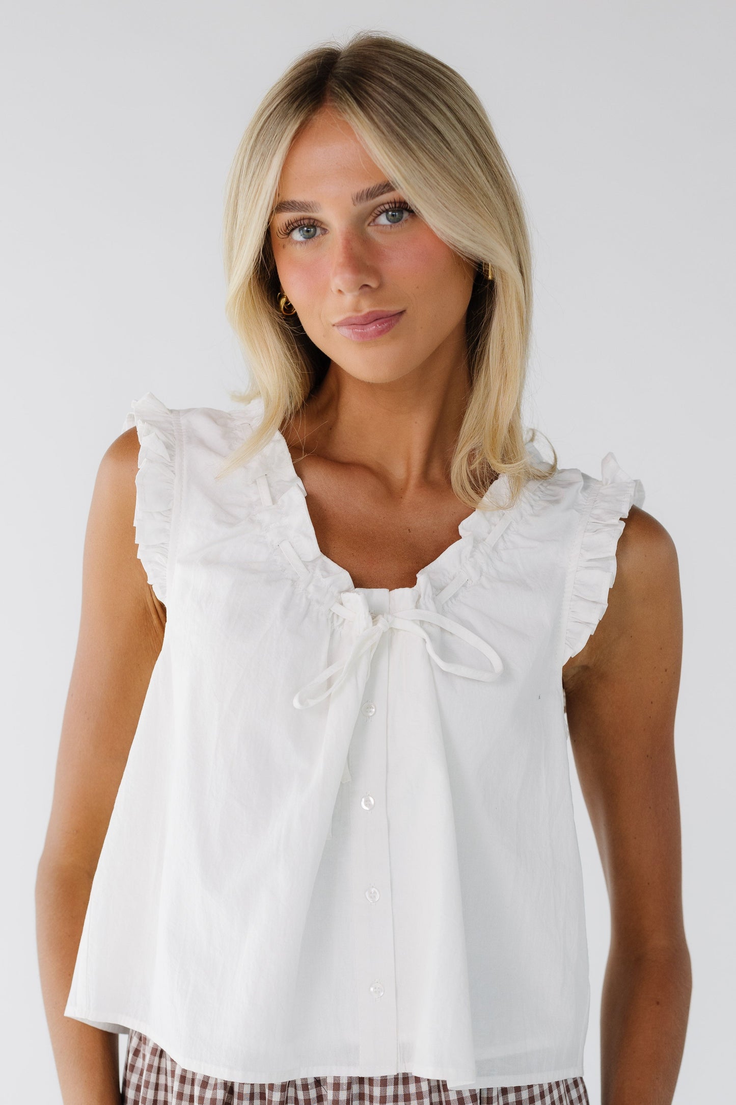Young woman wearing a white sleeveless button down blouse with ruffled armholes and a scoop ruffled neckline with a tie.