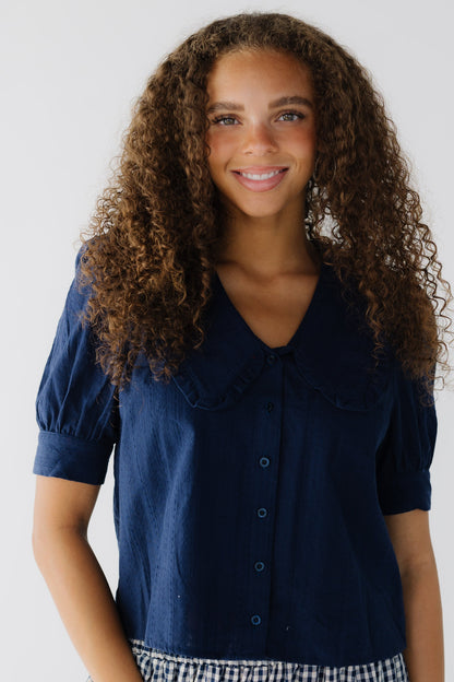 Teenage girl wearing a modest navy button down, textured blouse with a V-neckline and ruffled edge lapels, with cuffed short puff sleeves.