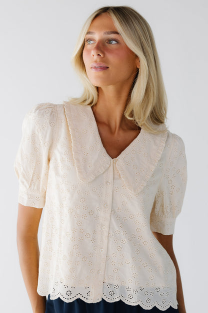 Young woman wearing a modest all eyelet cream blouse that is fully lined. It is a button down blouse with a scallop hem, short cuffed sleeves and a V-neckline with wide lapels.