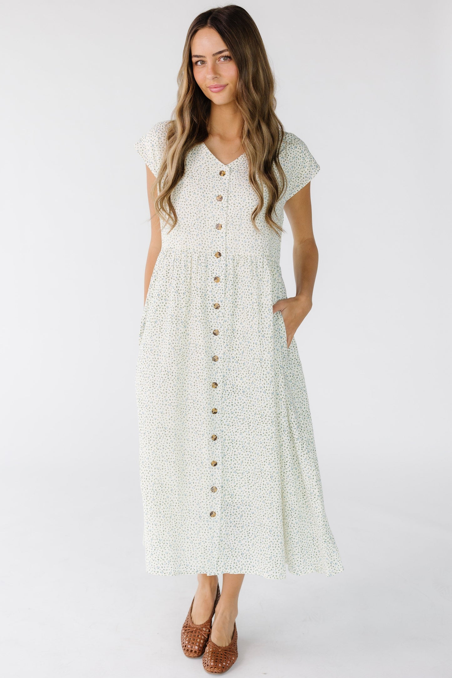 Young woman wearing a loose fitting midi dress in a white floral print that buttons down the front with side pockets, V-neckline and short sleeves.
