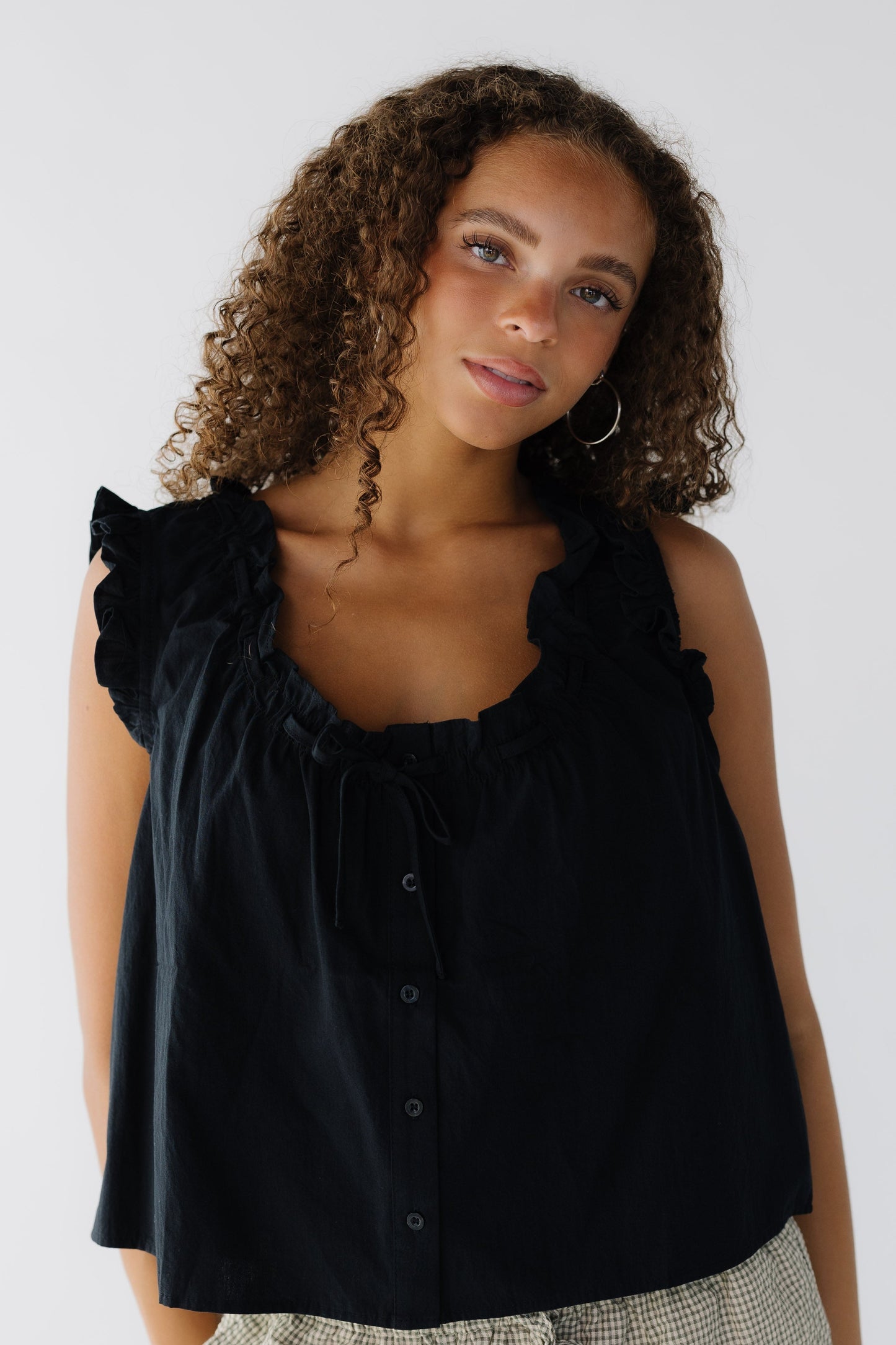 Young woman wearing a black loose fitting button down sleeveless blouse with ruffled armholes and a ruffled scoop neckline.