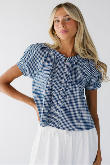 Young woman wearing a navy & white gingham button down blouse with front pleats, puff sleeves and a round neckline.