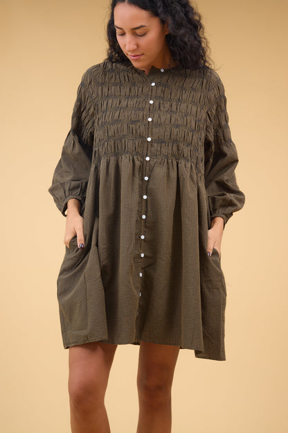 Young woman wearing a short brown & black gingahm button down dress with long sleeves, spaced out smocked bodice and side pockets.